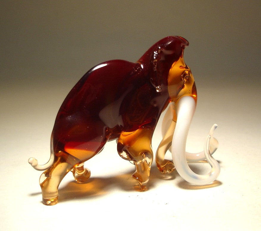 A detailed handmade glass mammoth figurine with long, curved tusks, depicted in a walking position.