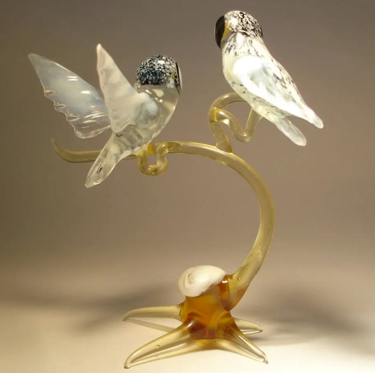 Side angle of a glass figurine showcasing two polar owls on a branch, one has its wings open. The owls are sitting together on an  elegantly curved branch.