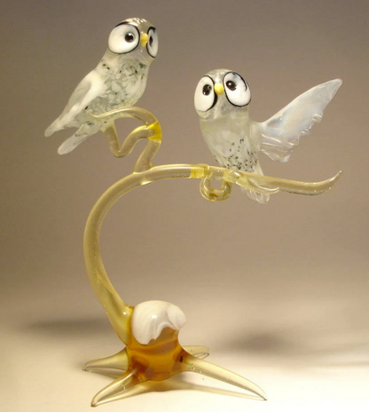 A close-up view of two glass polar owls perched on a branch. The owls are intricately designed with translucent white glass, featuring detailed feathers.