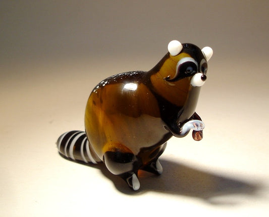 Close-up of the glass raccoon figurine, focusing on its expressive eyes, mask-like facial markings, and small paws.