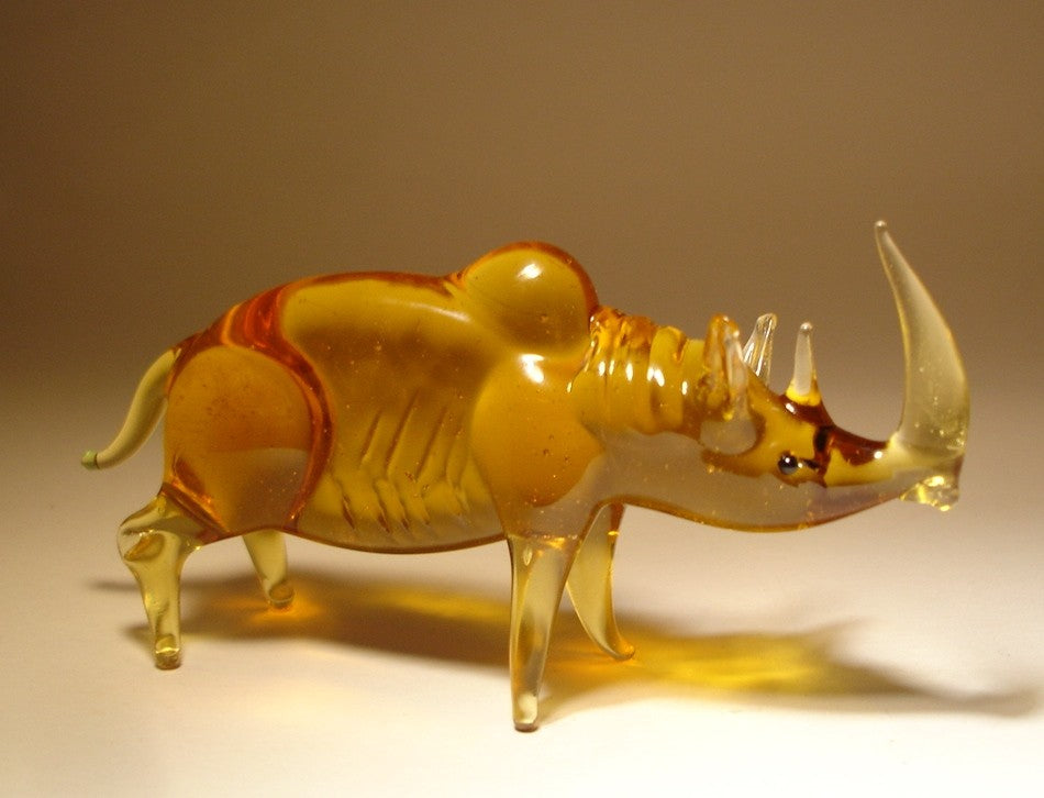 A side view of a glass rhinoceros figurine, highlighting its muscular build, curved horn, and intricate detailing along its back and legs.