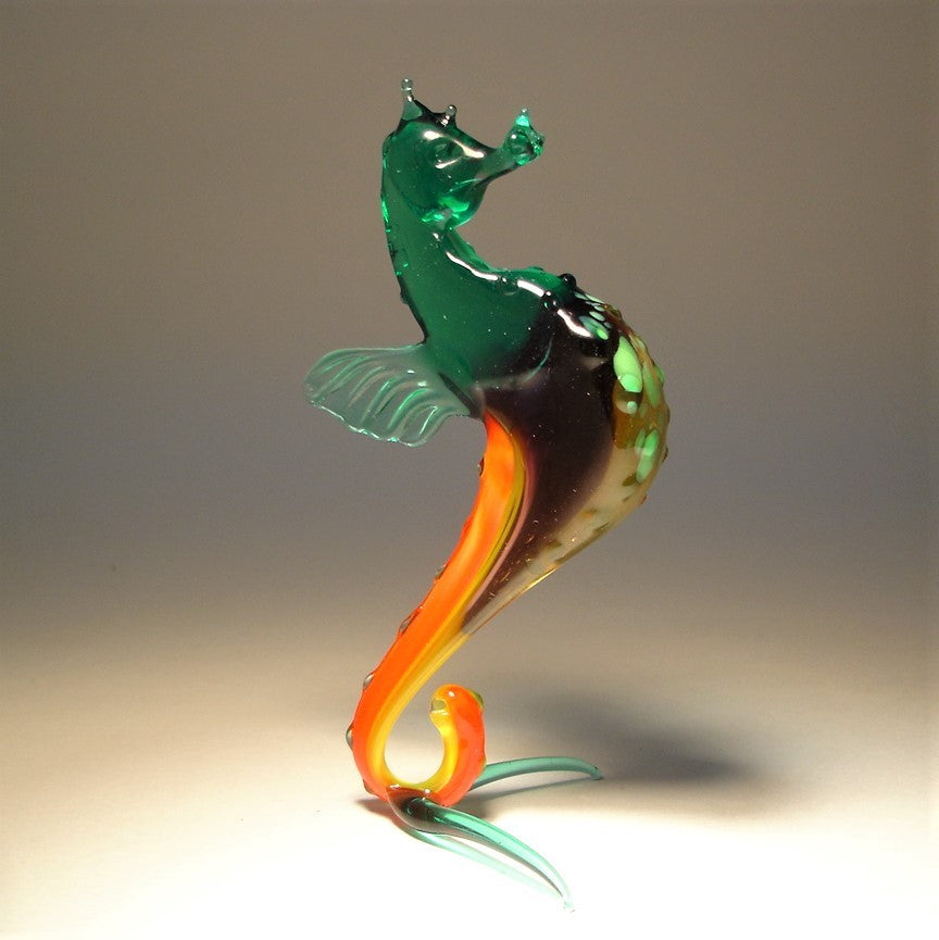 A glass figurine of a seahorse with a vibrant green body and orange accents, standing upright on its tail.