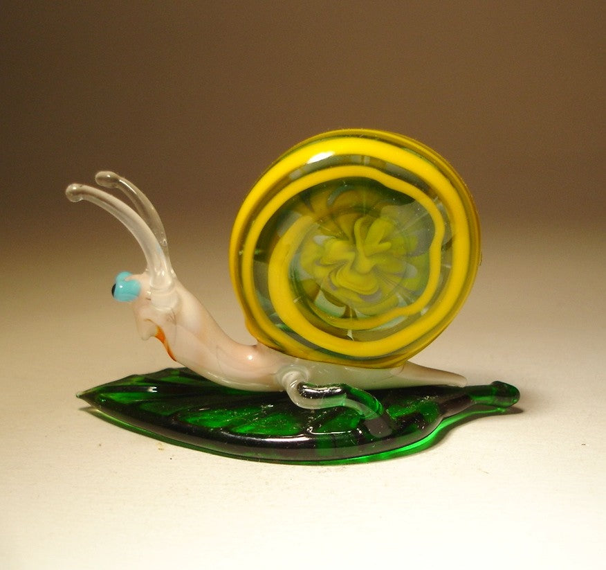 A top view of a glass snail figurine resting on a leaf, emphasizing the spiral shape of its shell and the rich green color of the leaf.