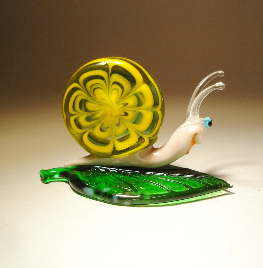 A front view of a glass snail figurine perched on a vibrant green leaf, showcasing its detailed yellow and green shell and delicate face features.