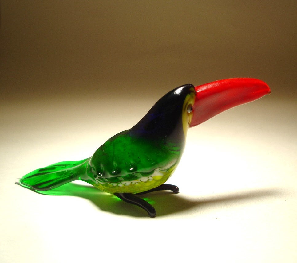 A side view of a glass toucan figurine, emphasizing the bold red beak and the smooth curves of its body.