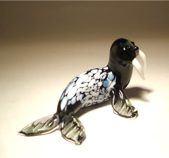 A side view of a glass walrus figurine, highlighting its sleek body with white spots, prominent tusks, and flippered limbs.