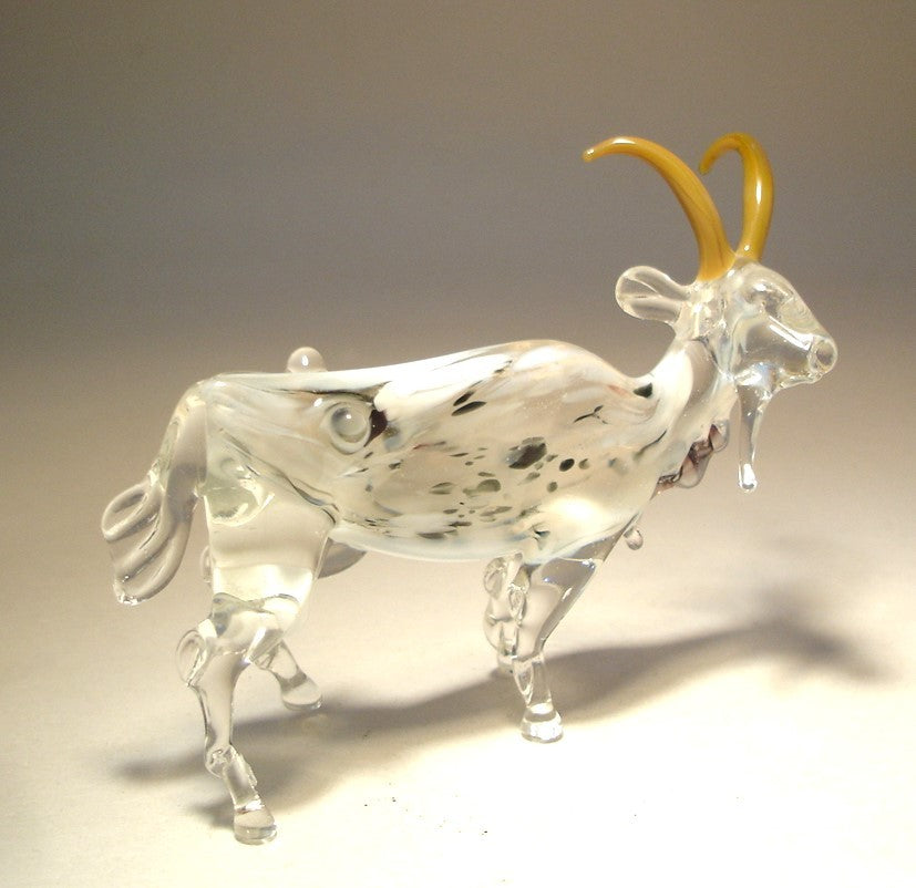 A side profile of the white and clear glass goat figurine, featuring the black and white markings on its body and light brown curved horns.