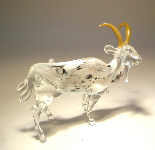 A side profile of the white and clear glass goat figurine, featuring the black and white markings on its body and light brown curved horns.