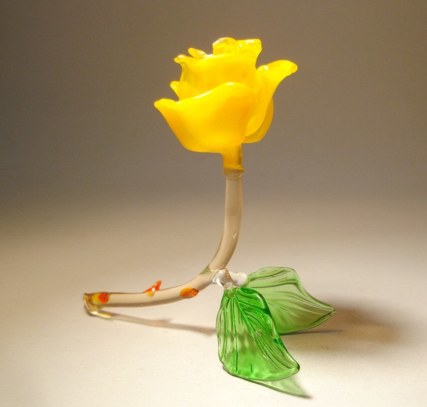 A side view of the yellow rose glass figurine, highlighting the layered petals as they gently open from the center. The transparent light brown stem curves naturally up, supporting the flower.